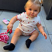 Cassie a rejoint le concours — aidez-le/la à gagner de superbes lots ! toddler, child, pigtails, strawberry_pattern, white_clothing, black_socks, toy, red_and_white_toy, outdoor, wicker_furniture, blue_towel, tile_floor, curious_expression, sitting, baby, person, cute, playful, daylight, portrait
