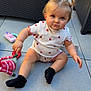toddler, child, pigtails, strawberry_pattern, white_clothing, black_socks, toy, red_and_white_toy, outdoor, wicker_furniture, blue_towel, tile_floor, curious_expression, sitting, baby, person, cute, playful, daylight, portrait