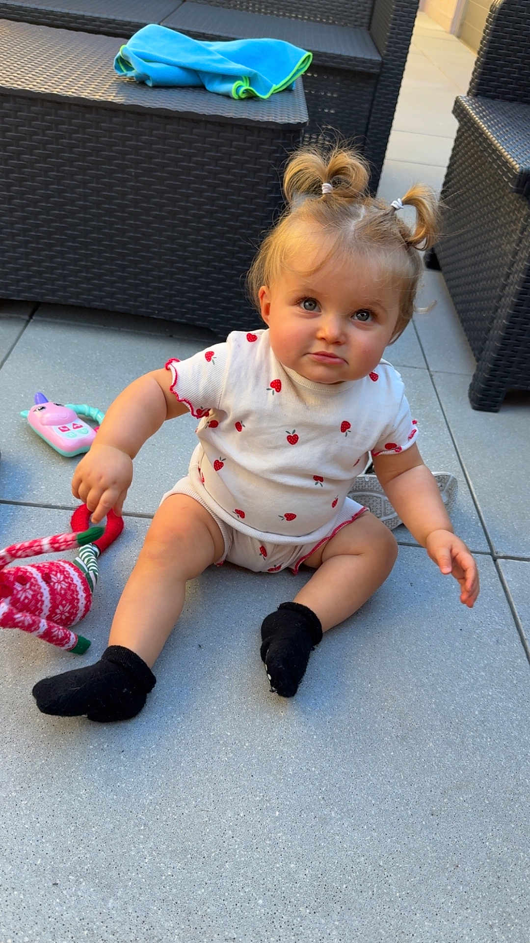 Cassie a rejoint le concours — aidez-le/la à gagner de superbes lots ! toddler, child, pigtails, strawberry_pattern, white_clothing, black_socks, toy, red_and_white_toy, outdoor, wicker_furniture, blue_towel, tile_floor, curious_expression, sitting, baby, person, cute, playful, daylight, portrait