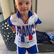 Tony participe au concours pour gagner de l'argent avec cette photo : child, boy, smiling, indoors, sporty, football, clothing, shorts, sneakers, laundry_basket, folded_clothes, tile_floor, short_hair, standing, casual, happy, portrait, home, young, cute