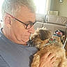 man, dog, glasses, gray_hair, indoor, couch, window, light, pet, affection, casual_clothing, portrait, hand, fur, collar, beard, living_room, comfort, close_up, relaxation