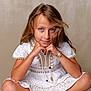child, girl, blue_eyes, white_dress, cross_legged, sandals, beaded_sandals, bracelet, long_hair, portrait, studio, smiling, cute, fashion, hands_under_chin, natural_light, casual_pose, indoor, young, innocent