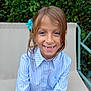child, smiling, blue_flower, striped_shirt, outdoor, chair, greenery, portrait, happy, casual_clothing, person, young, natural_light, relaxed, seated, blue_eyes, long_hair, buttoned_shirt, nature, daylight
