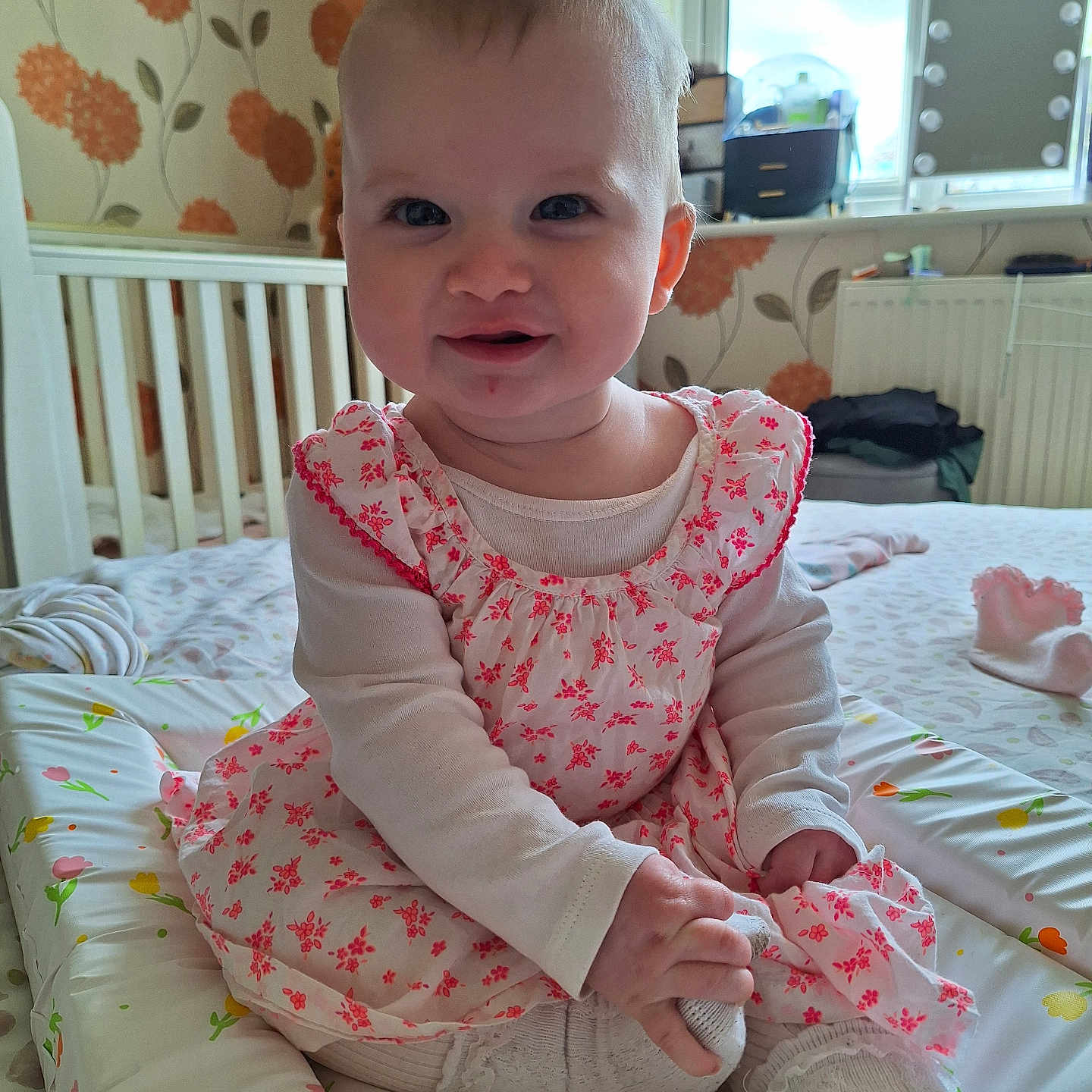 Rosie is registered to the contest to win money with this photo: baby, changing_mat, child, crib, cute, dress, floral_pattern, happy, indoor, infant, long_sleeve, natural_light, person, room, sitting, smiling, tights, toddler, wallpaper, window