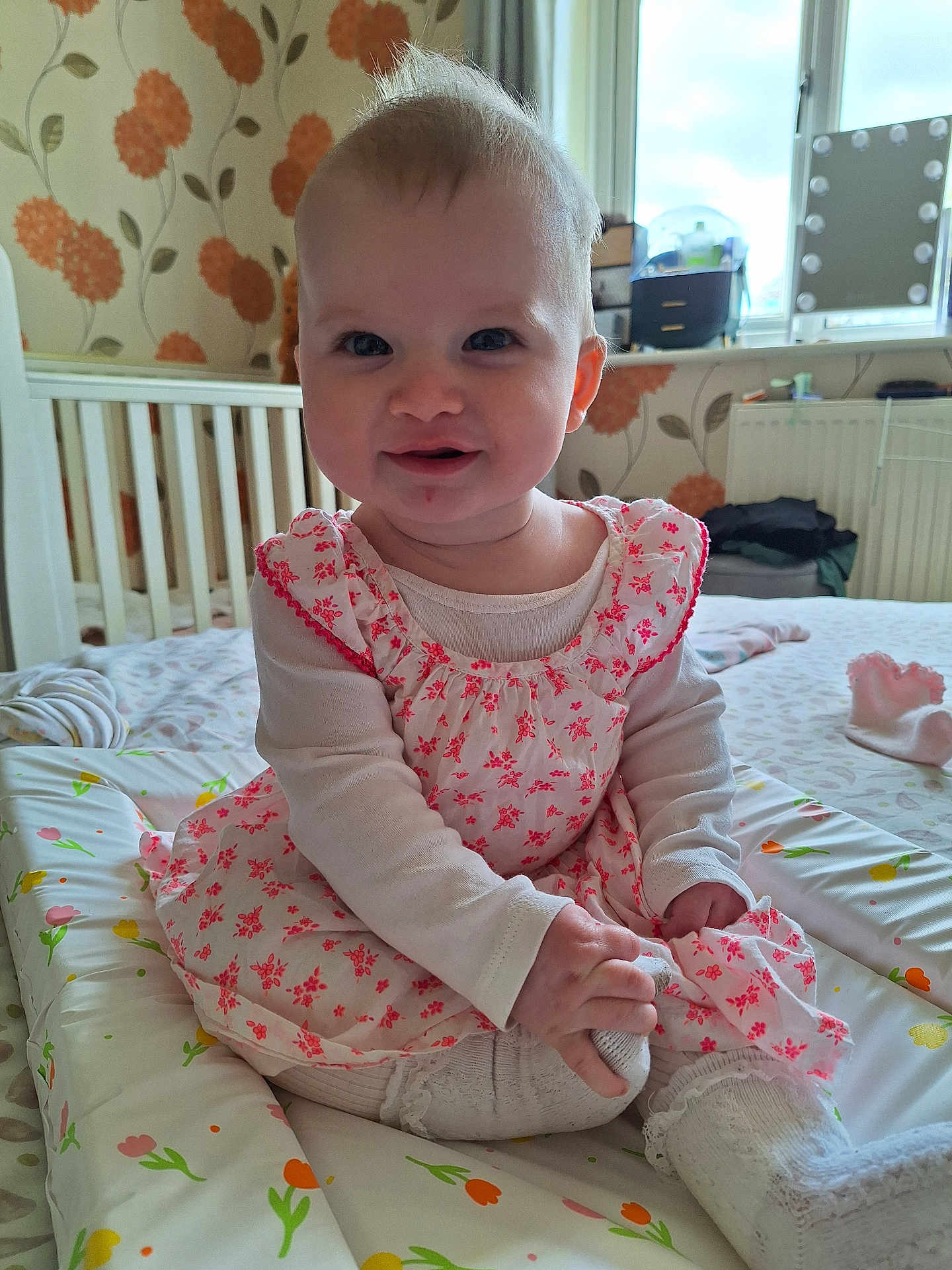 Rosie is registered to the contest to win money with this photo: baby, child, smiling, sitting, dress, floral_pattern, long_sleeve, tights, indoor, crib, wallpaper, window, natural_light, changing_mat, person, cute, happy, room, toddler, infant