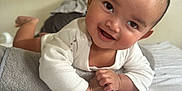 Aylann participe au concours pour gagner de l'argent avec cette photo : baby, barefoot, bed, blanket, cheerful, closeup, eyes, face, gaze, hair, hand, infant, lying_down, natural_light, pillow, portrait, smile, soft_texture, tummy_time, white_onesie