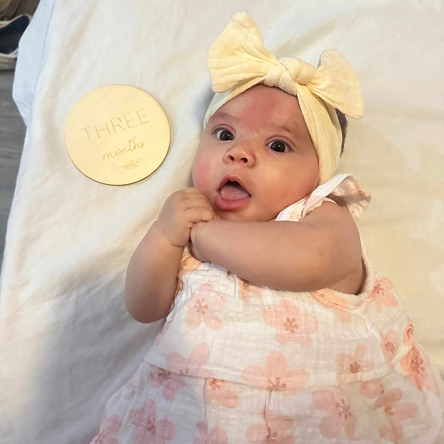 Evelynn joined the competition — help win amazing prizes! baby, blanket, bow, child, cute, dress, expression, eyes, face, floral, hand, headband, indoors, infant, lying_down, mouth, person, plaque, skin, wooden