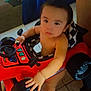 toddler, child, toy_car, indoor, floor_tile, steering_wheel, hand, barefoot, seat, checkered_pattern, red_color, black_color, playtime, curious, expression, person, young_child, race_car, baby, vehicle