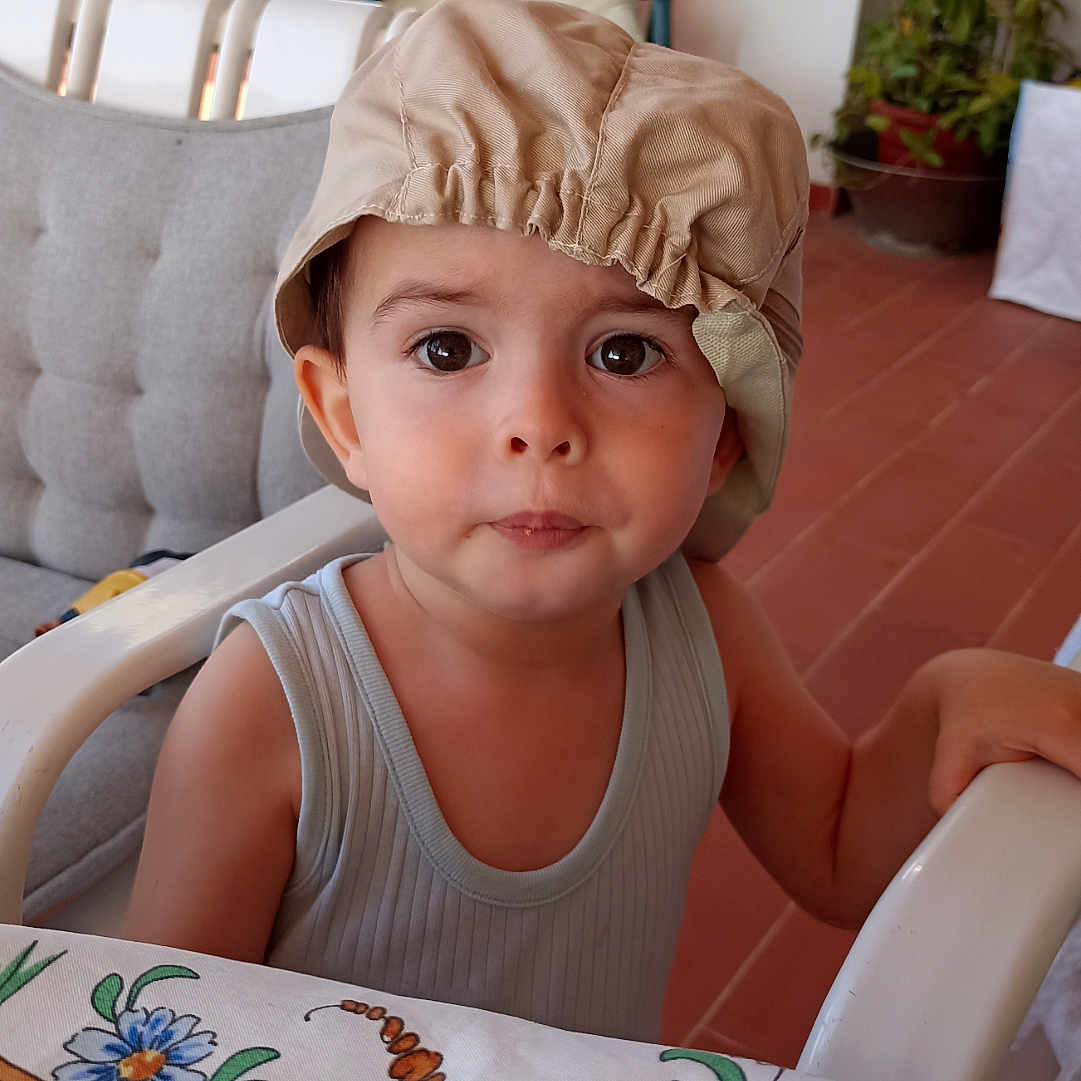 Joachim a rejoint le concours — aidez-le/la à gagner de superbes lots ! arm, casual_clothing, chair, child, curious, daylight, expression, eyes, face, floral_pattern, furniture, hat, home, indoor, person, plant, seated, table, tablecloth, toddler