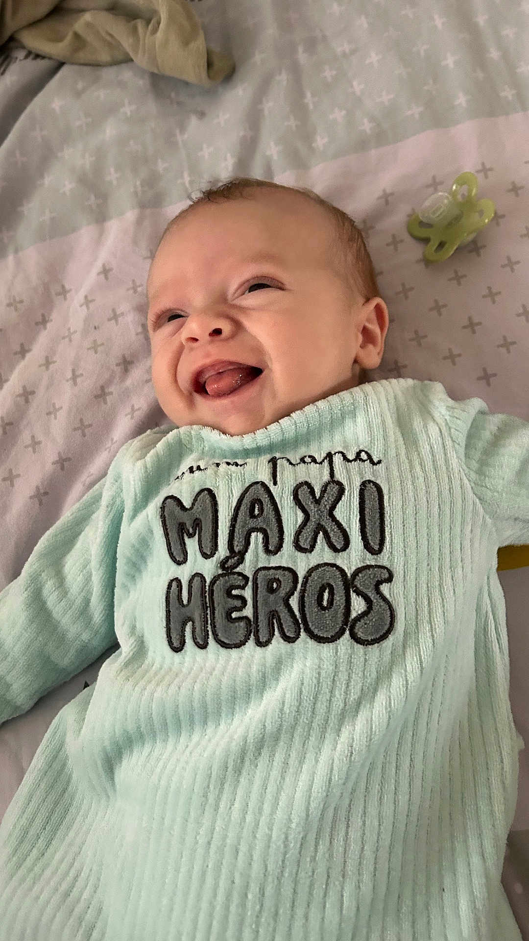 Charly participe au concours pour gagner de l'argent avec cette photo : baby, infant, smiling, onesie, pacifier, bed, blanket, face, mouth_open, tongue, happy, portrait, close_up, fabric, text_on_clothing, pattern, head, skin, nursery_bedding, pastel_color