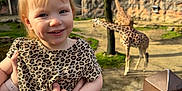 Ava is registered to the contest to win money with this photo: adult_hand, animal, child, daylight, fence, giraffe, grass, happy, leopard_print, nature, outdoor, person, pigtails, portrait, railing, shirt, smiling, toddler, tree, zoo