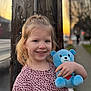 child, smiling, teddy_bear, plush_toy, outdoor, sunset, telephone_pole, wooden_pole, sidewalk, street, patterned_shirt, pink_shirt, portrait, happy, blond_hair, hand, bokeh, urban, person, photography