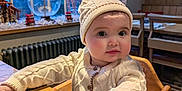 Bowen Belle joined the competition — help win amazing prizes! baby, child, high_chair, sweater, hat, wooden_table, indoor, lamp, window, cozy, cute, warm_lighting, furniture, hands, face, curious, decorations, winter_clothing, seated, portrait