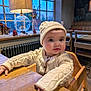 baby, child, high_chair, sweater, hat, wooden_table, indoor, lamp, window, cozy, cute, warm_lighting, furniture, hands, face, curious, decorations, winter_clothing, seated, portrait