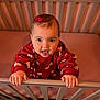 baby, crib, red_clothing, bow, happy, child, indoor, infant, cute, smiling, holding, railing, person, home, toddler, playful, portrait, small_hands, bedroom, cheerful