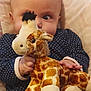 baby, child, plush_toy, giraffe, stuffed_animal, polka_dot_clothing, hand, face, eyes, blanket, infant, cute, soft_toy, lying_down, cozy, indoor, portrait, young_child, close_up, curious