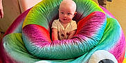 Malachi is registered to the contest to win money with this photo: baby, infant, bean_bag, colorful, rainbow, poop_emoji, indoor, person, smiling, plush, furniture, child, cute, seat, toy, soft, candid, happy, home, googly_eyes