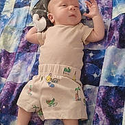 Xavion is registered to the contest to win money with this photo: baby, infant, child, quilt, blanket, patterned_clothing, shorts, shirt, lying_down, hand_gesture, skin, face, foot, relaxed, indoors, colorful, purple, blue, white, cartoon