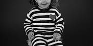 Nayla participe au concours pour gagner de l'argent avec cette photo : child, girl, portrait, black_and_white, striped_clothing, knitwear, heart_patch, curly_hair, bow, sitting, stool, sandals, toes, studio, smiling, cute, fashion, young_child, innocence, monochrome