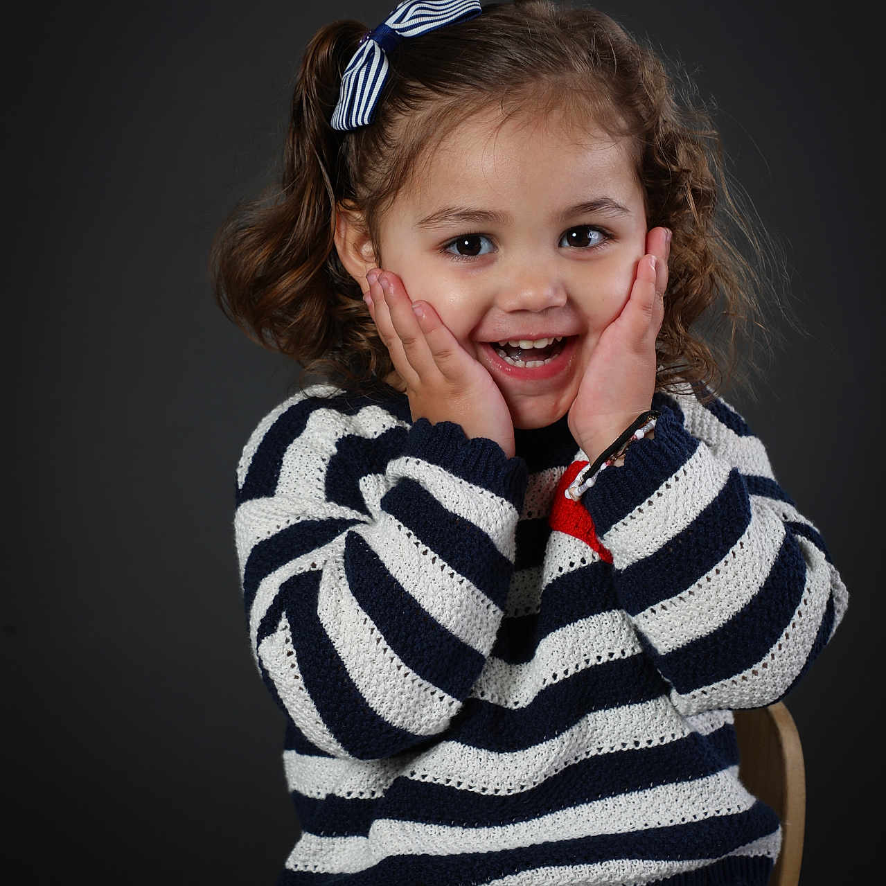 Nayla a rejoint le concours — aidez-le/la à gagner de superbes lots ! toddler, child, smiling, happy, portrait, striped_clothing, knitwear, bow, curly_hair, seated, studio, dark_background, cute, hands_on_cheeks, person, fashion, casual, young_child, expression, indoor