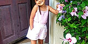 Sabi participe au concours pour gagner de l'argent avec cette photo : child, girl, flower, door, outdoor, plant, greenery, pink_clothing, shorts, sandals, sidewalk, posing, smiling, nature, home, step, wall, purple_flower, sunlight, casual