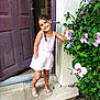 child, girl, flower, door, outdoor, plant, greenery, pink_clothing, shorts, sandals, sidewalk, posing, smiling, nature, home, step, wall, purple_flower, sunlight, casual