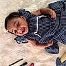 baby, child, smiling, blue_dress, striped_clothing, flower_hair_accessory, lying_down, white_fur_rug, pearl_necklace, straw_hat, makeup_items, happy, cute, portrait, indoor, person, infant, accessories, fashion, playful