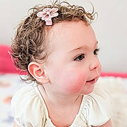 Kira is registered to the contest to win money with this photo: baby, toddler, child, curly_hair, hair_clip, flower_clip, profile, rosy_cheeks, ear, eyelashes, white_dress, ruffle, portrait, cute, soft_light, indoors, bedroom, skin, smile, closeup