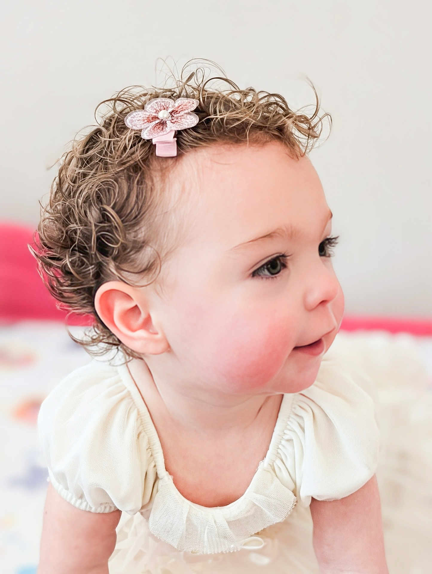 Kira is registered to the contest to win money with this photo: baby, toddler, child, curly_hair, hair_clip, flower_clip, profile, rosy_cheeks, ear, eyelashes, white_dress, ruffle, portrait, cute, soft_light, indoors, bedroom, skin, smile, closeup