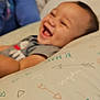 Mateo is registered to the contest to win money with this photo: arrows, baby, bedroom, blue_shirt, blurred_background, candid, child, closeup, clothing, decor, happy, indoor, infant, laughing, lying_down, person, pillow, smile, soft_lighting, text