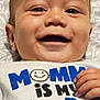 baby, child, closeup, clothing, cute, dimples, face, hand, happy, indoors, infant, joyful, patterned_background, person, portrait, smiley_face, smiling, text, white_shirt, young