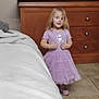 child, girl, purple_dress, bedroom, dresser, television, floor_tile, bed, blanket, furniture, indoor, person, standing, young_child, hair, clothing, toy_cup, plastic_bottle, drawer, smile