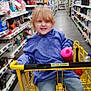 child, shopping_cart, store_aisle, red_hair, blue_sweater, jeans, pink_cup, smile, indoor, retail, products, shelves, floor, yellow_cart, casual_clothing, young_child, playful, holding, grocery_store, person