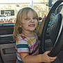 child, toddler, car, steering_wheel, smile, lollipop, sitting, window, daylight, interior, clothing, shorts, shirt, seat, happy, blond_hair, face, hand, footwear, vehicle