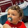 toddler, child, pacifier, red_shirt, curly_hair, indoor, dining_room, wine_glasses, table, chair, face, portrait, looking_away, young_child, person, casual_clothing, cute, baby, expression, seated