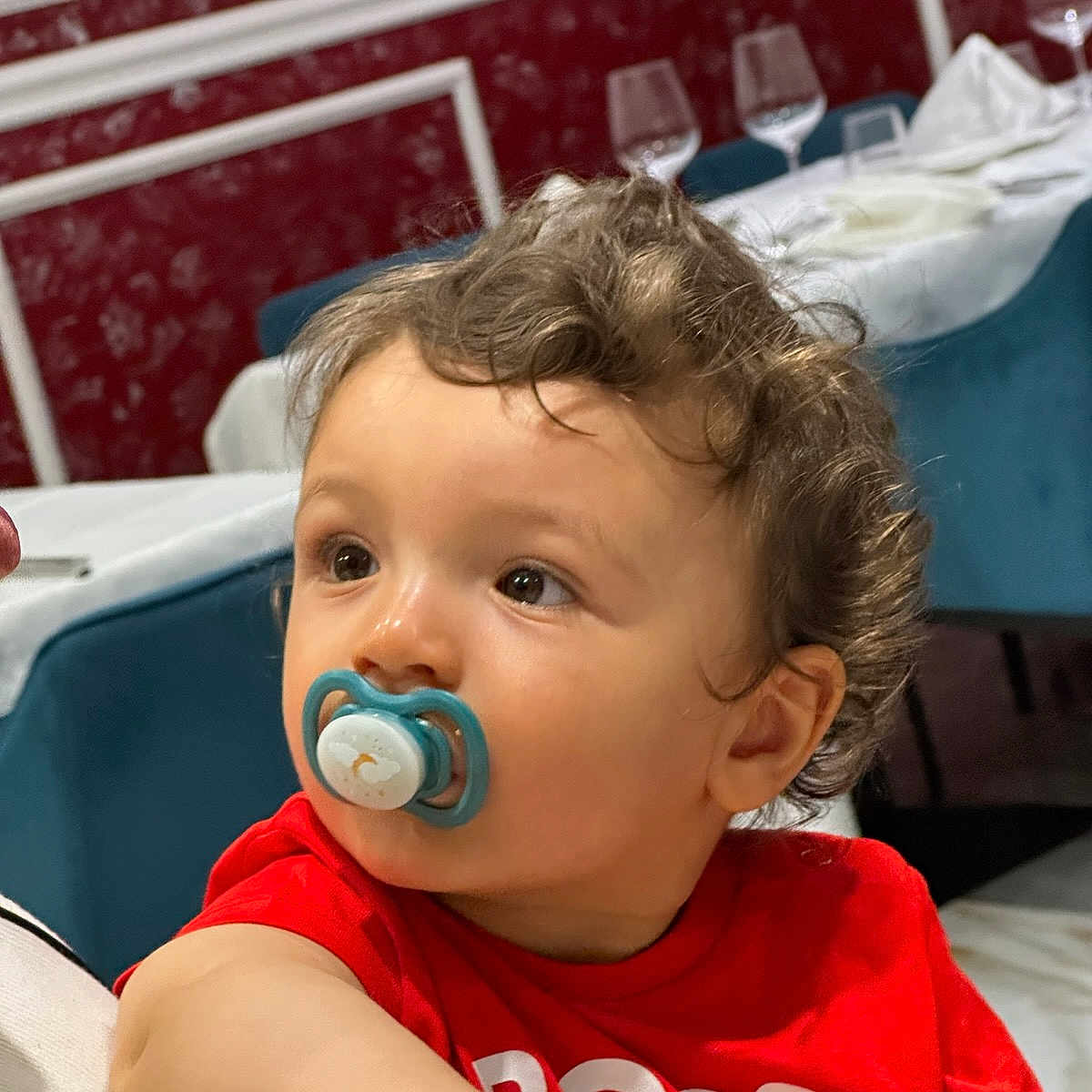 Ridha a rejoint le concours — aidez-le/la à gagner de superbes lots ! baby, casual_clothing, chair, child, curly_hair, cute, dining_room, expression, face, indoor, looking_away, pacifier, person, portrait, red_shirt, seated, table, toddler, wine_glasses, young_child