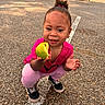 child, toddler, apple, bite, pink_clothing, hair_bun, sneakers, asphalt, parking_lot, cars, houses, outdoor, daylight, playful, happy, crouching, holding, food, smiling, person