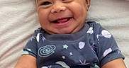 Sage is registered to the contest to win money with this photo: baby, smiling, child, infant, onesie, space_theme, blanket, bed, happy, cute, face, portrait, lying_down, skin, hair, indoors, person, toddler, clothing, cozy