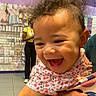 Sage is registered to the contest to win money with this photo: baby, child, smiling, happy, curly_hair, floral_clothing, person, indoor, store, retail, woman, yellow_top, accessories, earrings, holding, hand, floor_tiles, purple_wall, merchandise, shopping