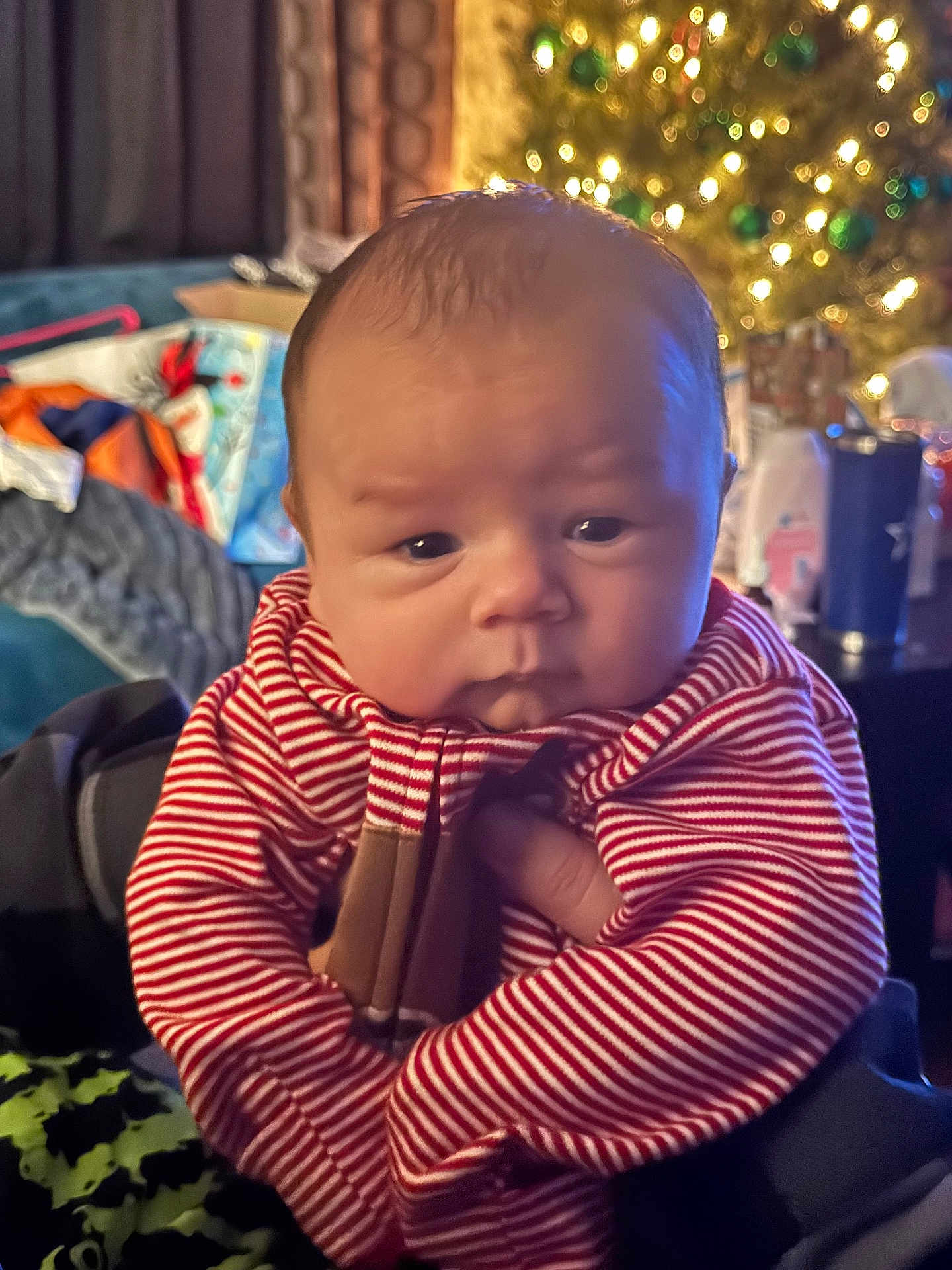 Dakota joined the competition — help win amazing prizes! baby, child, striped_clothing, face, indoor, christmas_tree, holiday_lights, hand, person, warm_lighting, cozy, infant, portrait, cute, newborn, soft_focus, background_blur, furniture, holiday_decor, smile