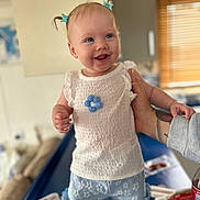 Laney joined the competition — help win amazing prizes! baby, child, smiling, blue_hair_clips, white_top, blue_shorts, flower_pattern, tattooed_arm, indoor, table, bottle, monitor, window_blinds, happy, person, cute, standing, supporting_hand, clothing, portrait