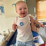 baby, child, smiling, blue_hair_clips, white_top, blue_shorts, flower_pattern, tattooed_arm, indoor, table, bottle, monitor, window_blinds, happy, person, cute, standing, supporting_hand, clothing, portrait