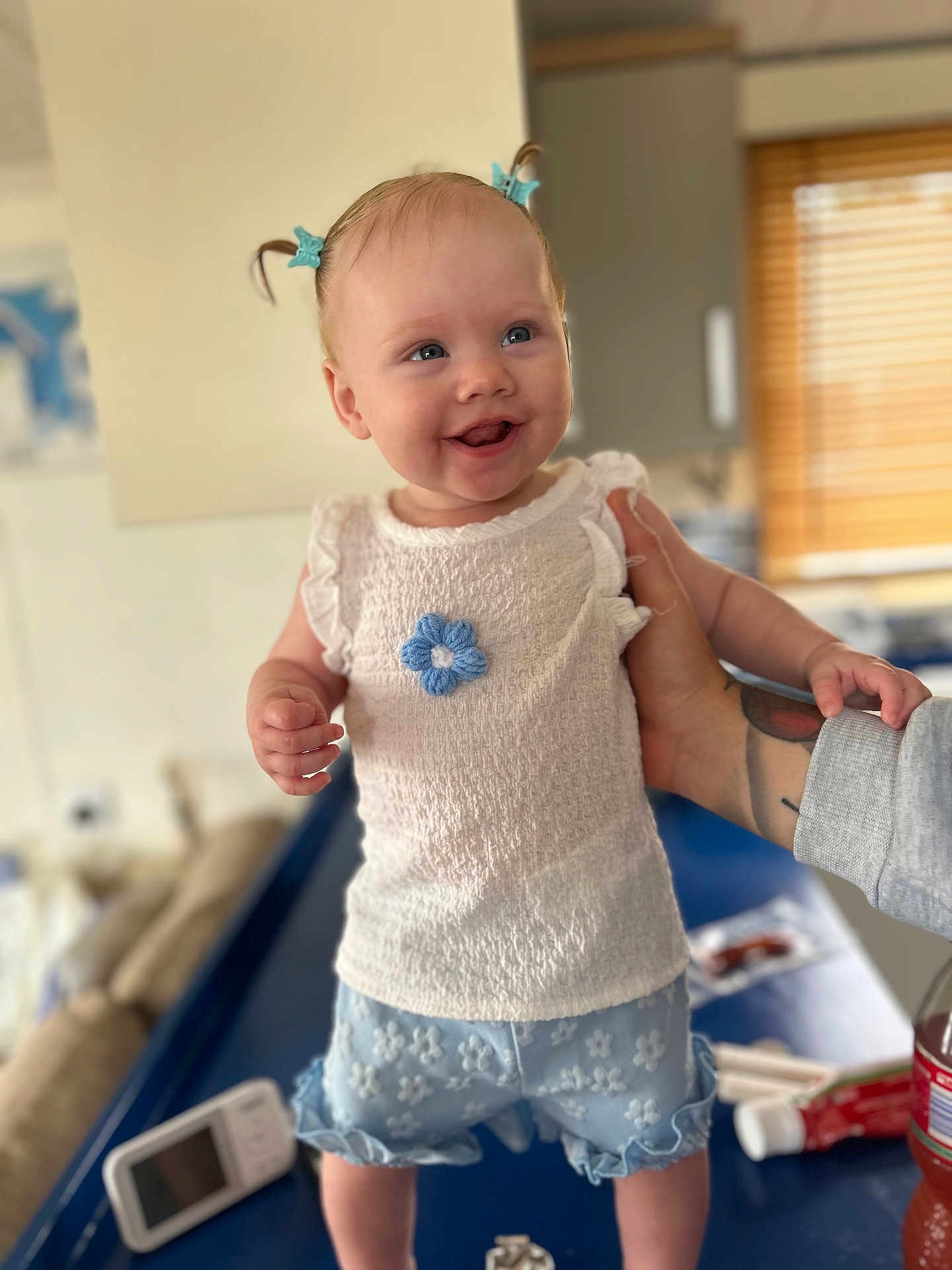 Laney joined the competition — help win amazing prizes! baby, child, smiling, blue_hair_clips, white_top, blue_shorts, flower_pattern, tattooed_arm, indoor, table, bottle, monitor, window_blinds, happy, person, cute, standing, supporting_hand, clothing, portrait