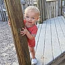 toddler, child, playground, wooden_post, red_striped_clothing, white_shoes, outdoor, daylight, fence, wood, gravel, curious, peeking, young_child, person, smiling, standing, nature, summer, casual