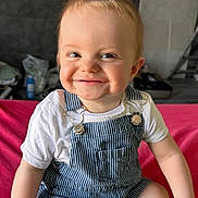 Jules participe au concours pour gagner de l'argent avec cette photo : toddler, child, smiling, overalls, white_shirt, sitting, red_surface, indoor, face, happy, cute, baby, person, clothing, shorts, skin, hair, hand, legs, portrait