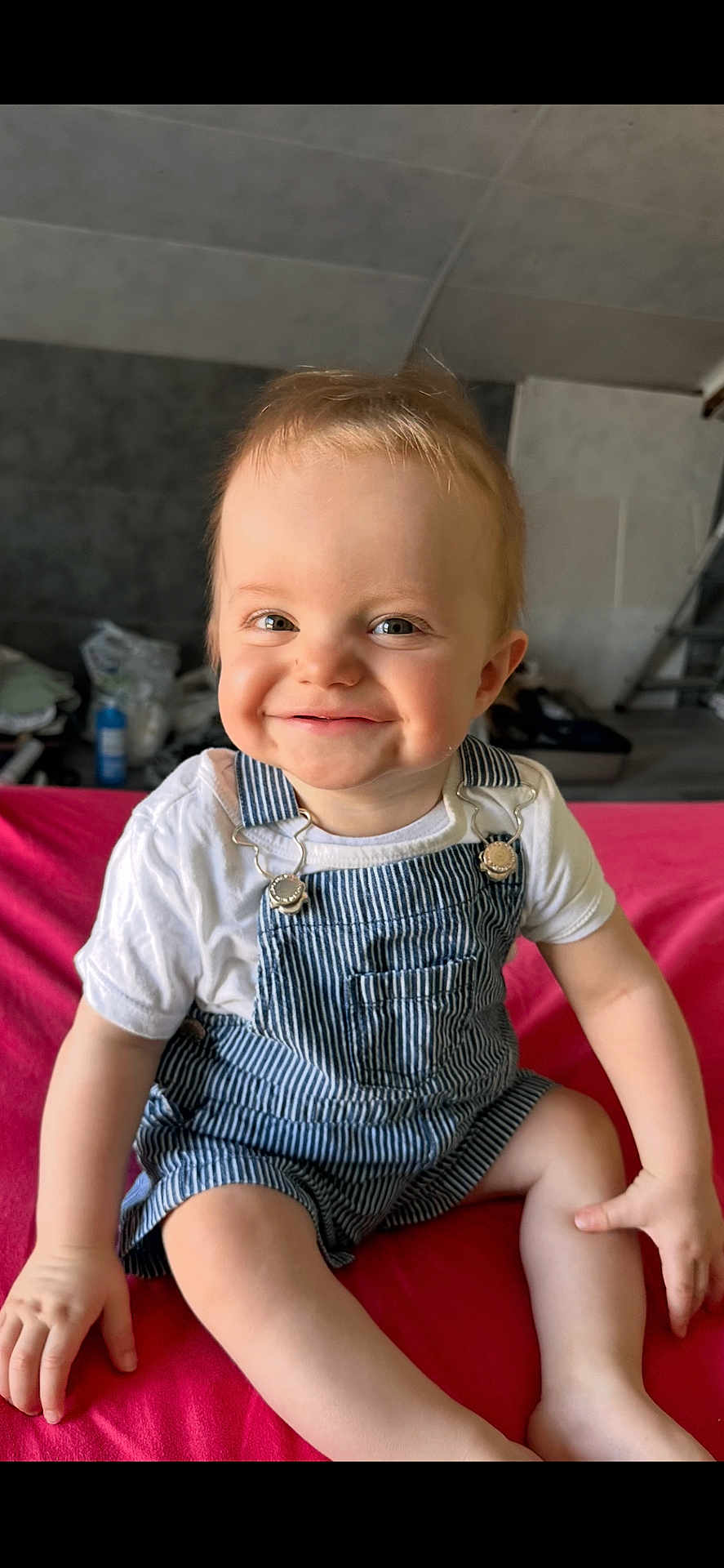 Jules participe au concours pour gagner de l'argent avec cette photo : toddler, child, smiling, overalls, white_shirt, sitting, red_surface, indoor, face, happy, cute, baby, person, clothing, shorts, skin, hair, hand, legs, portrait