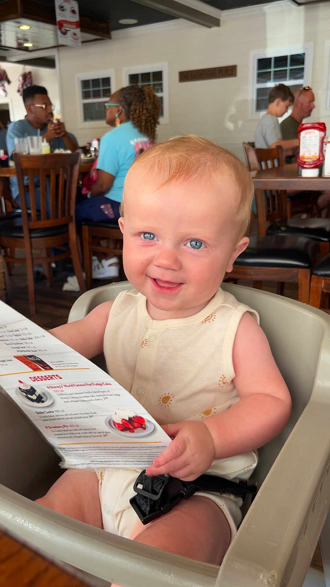 Weston is registered to the contest to win money with this photo: baby, high_chair, menu, restaurant, smile, child, indoor, seat, table, people, food_menu, happy, curly_hair, blue_eyes, casual_clothing, dining, family, childhood, candid, portrait