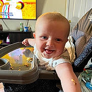 Weston is registered to the contest to win money with this photo: baby, high_chair, smiling, food, crumbs, arm, television, cartoon, indoor, child, person, furniture, happy, reaching, clothing, face, table, room, home, seat