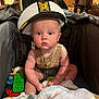 Weston is registered to the contest to win money with this photo: baby, child, captain_hat, titanic, playpen, toy, blanket, indoor, curious, drool, sitting, clothing, person, face, headwear, furniture, wooden_cabinet, plastic_toy, soft_toy, footwear