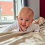 amber_necklace, baby, bed, blanket, blue_eyes, child, cozy, curtain, face, happy, head, indoor, infant, natural_light, person, room, skin, smiling, white_clothing, window